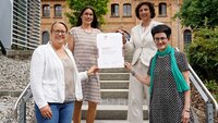 Vier Frauen stehen lächelnd auf einer Treppe vor einem Gebäude und halten ein Blatt Papier fest