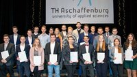 23 Studierende stehen in mehreren Reihen hintereinander mit ihren Zeugnissen in der Hand auf der Bühne vor einem Banner mit dem TH-Logo