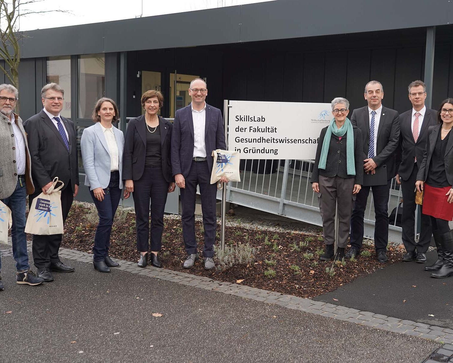 Gruppenbild mit 9 Personen vor dem Schild und dem Gebäude der Fakultät Gesundheitswissenschaften