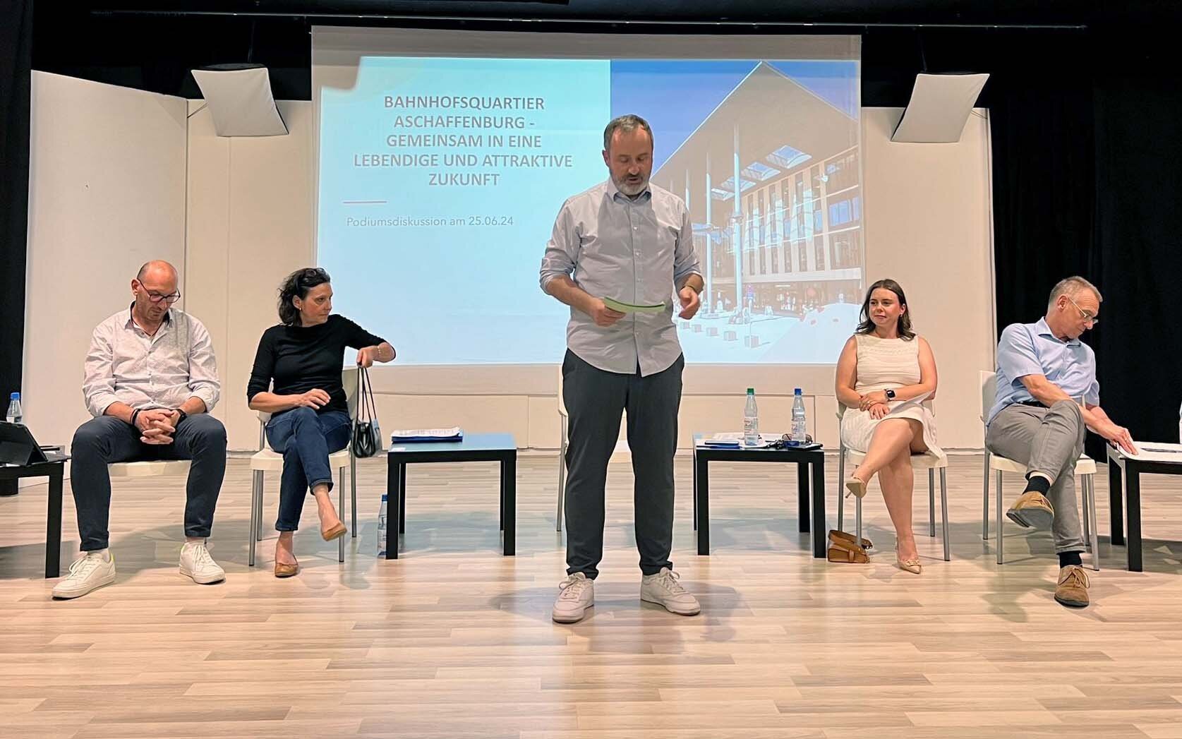 Der Moderator steht mit dem Rücken zu den Teilnehmenden der Podiumsdiskussion und lies etwas von einem Blatt Papier ab, das er in den Händen hält.