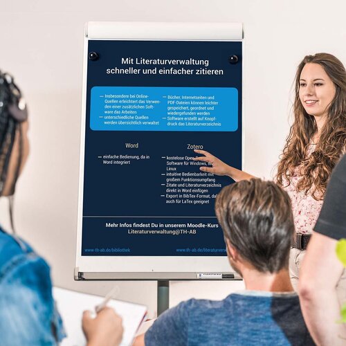 Drei Studierende hören einer Studierende, Referentin, Professorin, Dozentin zu, zeigt auf Flipchart mit Infos zum Zitieren