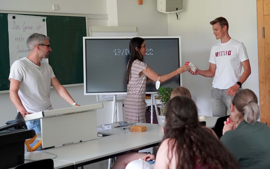 Eine Frau überreicht einem jungen Mann einen Gegenstand. Im Vordergrund sind einige Personen sitzend von hinten zu sehen.