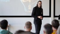 Ben Lenk-Ostendorf bei seinem Impulsvortrag mit Blick zum Publikum, im Hintergrund eine Glühbirne auf der Leinwand mit Überschrift Chain of thought