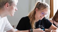 Ein Student und eine Studentin sitzen nebeneinander an einem Tisch und schauen gemeinsam in ein Buch.