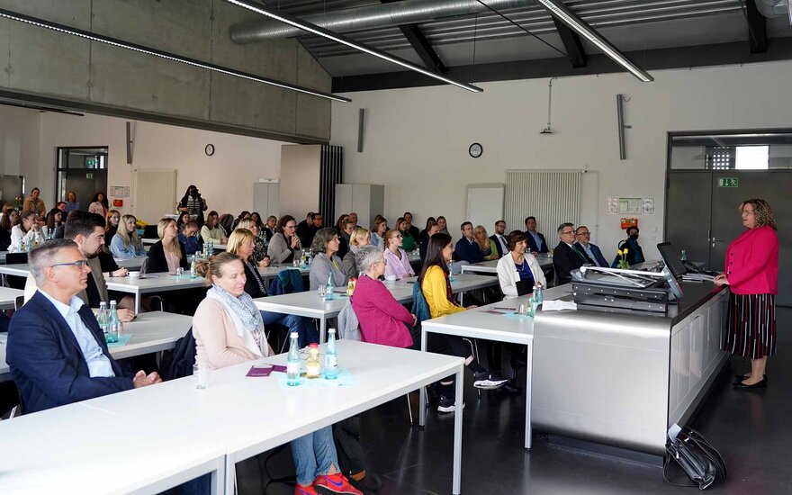 Eine Frau steht an einem breiten Tisch und spricht zum Publikum