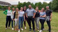 Eine Gruppe von neun Personen steht auf der grünen Campuswiese vor grünen Bäumen und dem Gebäude der Mensa bei leicht bewölktem Himmel