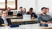 Studierende, die in mehreren Reihen hintereinander an Tischen in einem Hörsaal sitzen und lächelnd nach vorne blicken