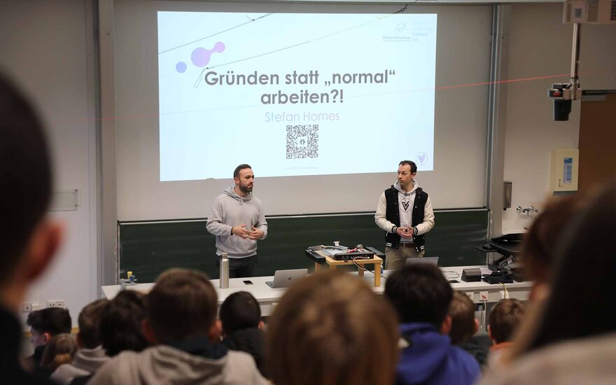 Zwei junge Männer sprechen vor einer Präsentationsleinwand zu den Schülerinnen und Schülern, die im Hörsaal sitzen