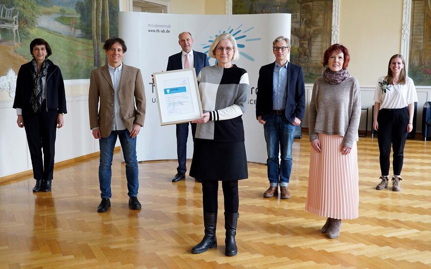 Eine Gruppe von 7 Personen, von denen eine eine Urkunde in der Hand hält, stehen vor der Wand mit dem Logo der Hochschule im Hock-Saal