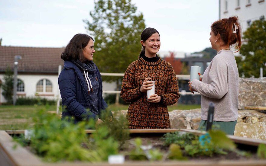 Drei Studentinnen unterhalten sich fröhlich lachend hinter einem Hochbeet mit Kräutern und Salat. Sie halten Kaffeebecher in der Hand.