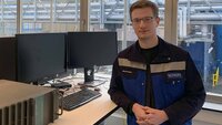 Ein junger Mann mit Brille im blauen Arbeitsanzug lehnt an einem Tisch mit Monitoren und technischen Geräten im Hintergrund