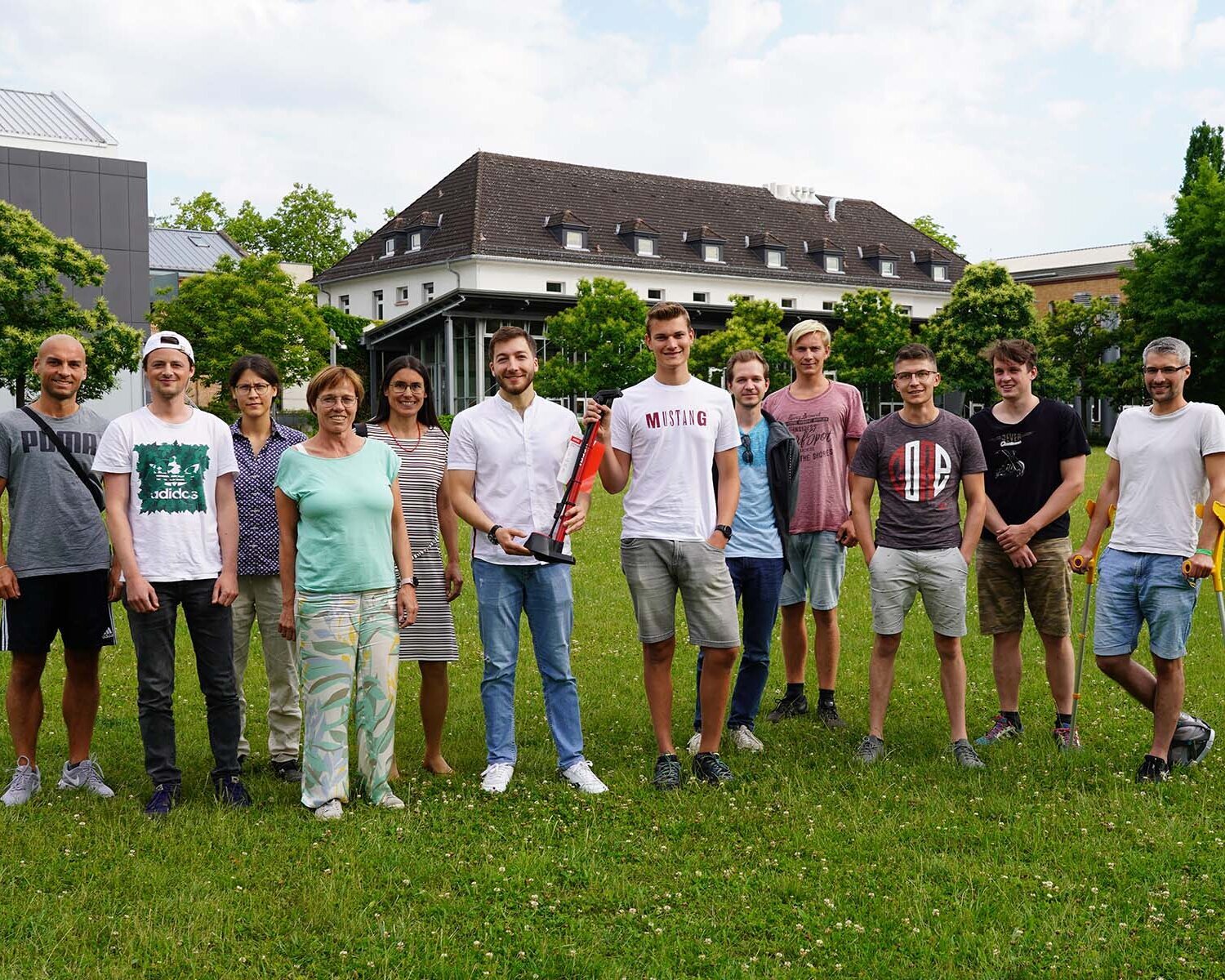 Gruppe von zwölf Personen steht auf einer grünen Wiese vor einem Gebäude und grünen Bäumen