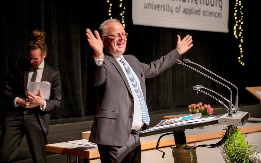 Ein grauhaariger Mann mit Brille steht mit ausgebreiteten Armen m Rednerpult vor einem Banner mit TH-Logo