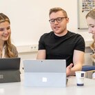 Luisa und zwei ihrer Kommilitonen sitzen vor zwei Laptops und unterhalten sich dabei lachend.