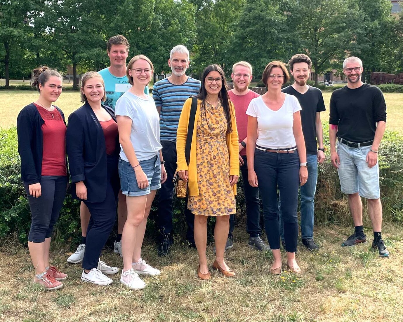 Gruppe von zehn Personen steht auf einer grünen Wiese, im Hintergrund grüne Bäume