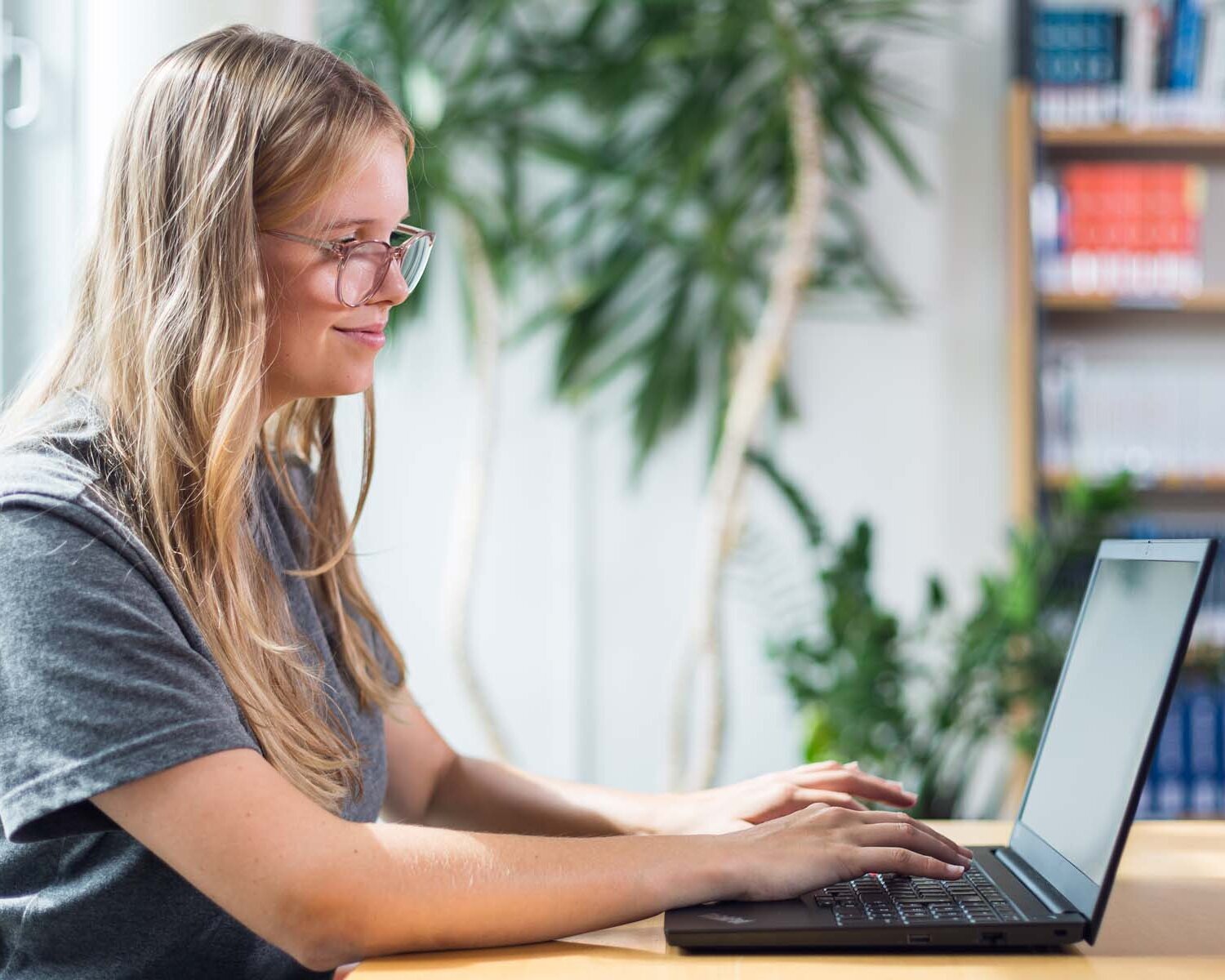 Blonde Frau sitzt an einem Tisch und arbeitet an einem schwarzen Laptop. Im Hintergund ist eine Pflanze und ein Bücherregal.