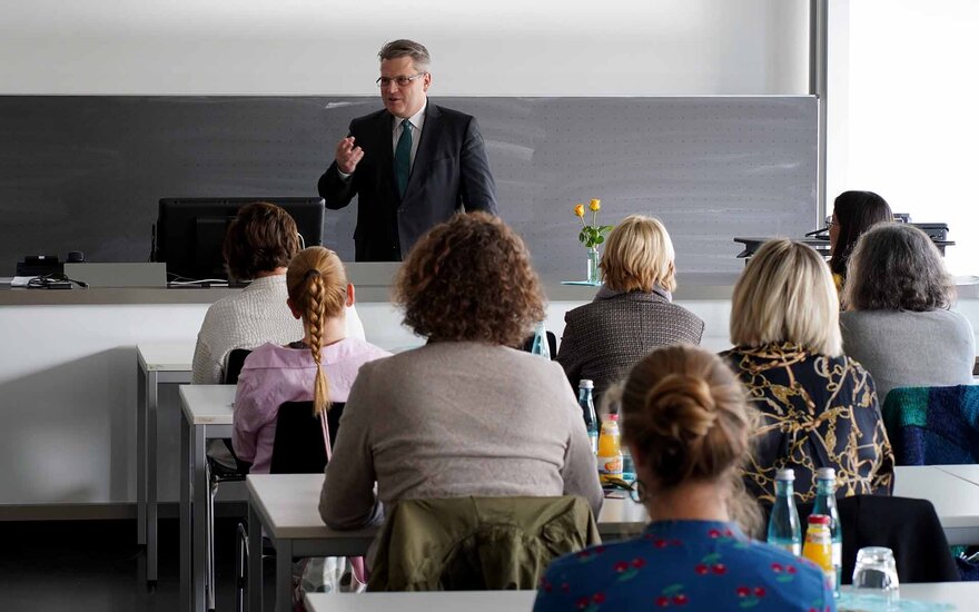 Eine Mann im dunklen Anzug steht an einem breiten Tisch und spricht zum Publikum