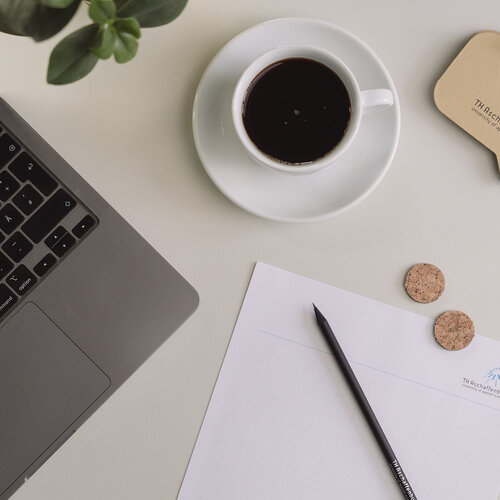 Auf einem Tisch steht ein Laptop, ein Kaffee, eine Pflanze, ein Post-It Block und Papier mit Stift.