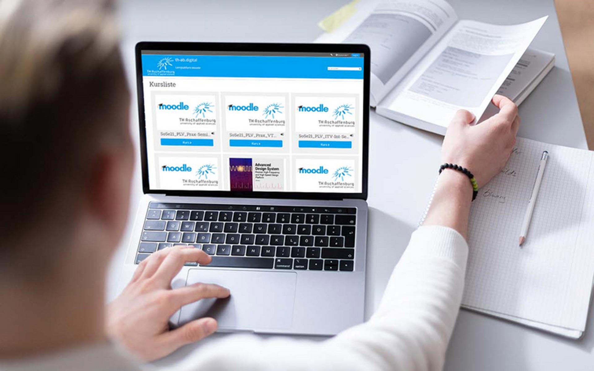 Ein Mann von hinten ist am Laptop und hat das Moodle der TH Aschaffenburg geöffnet. Mit der rechten Hand blättert er in einem Buch.