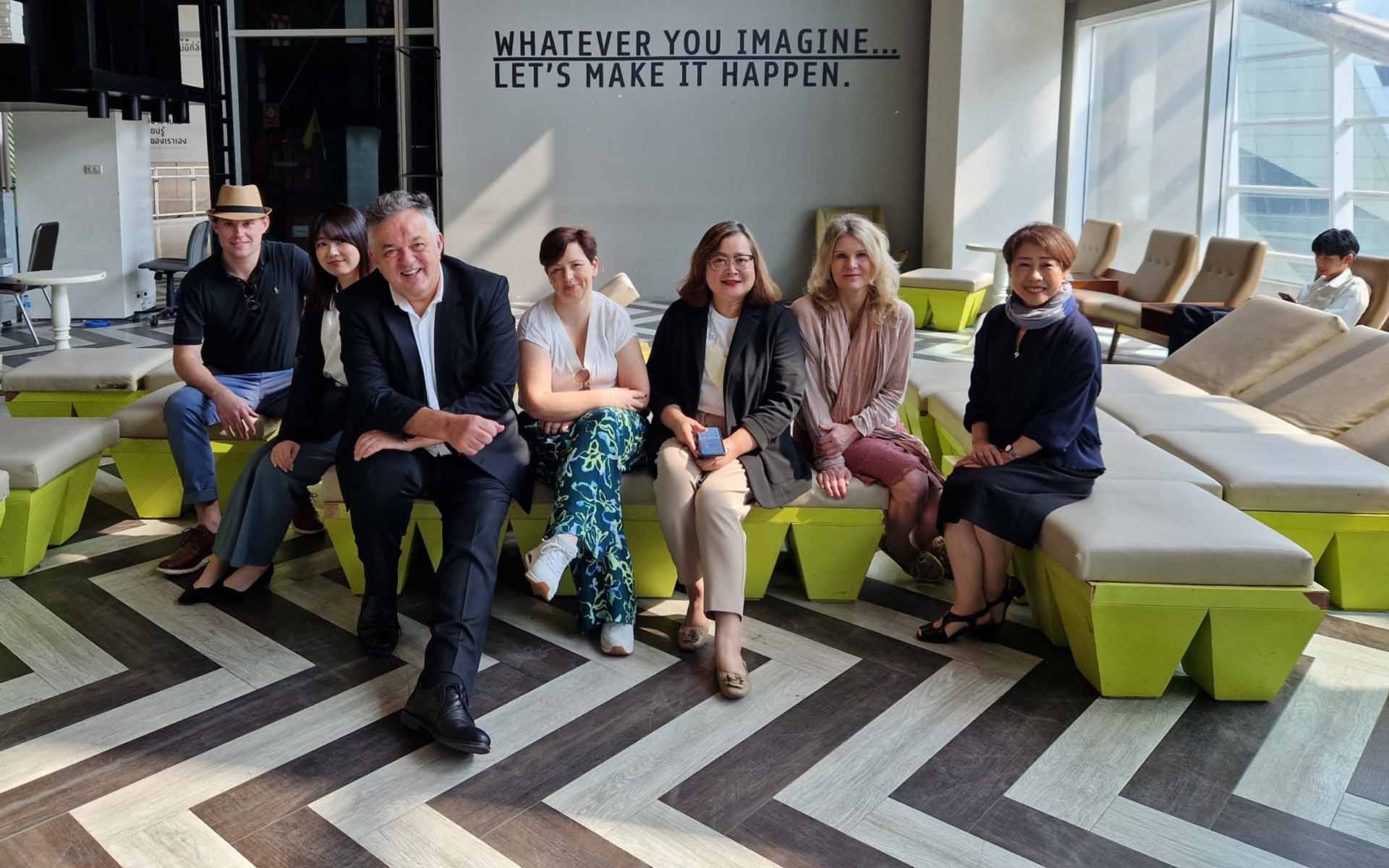 Eine Gruppe von sieben Personen sitzt gemeinsam in einer Loungeecke im Foyer eines Gebäudes. Alle lächeln in die Kamera.