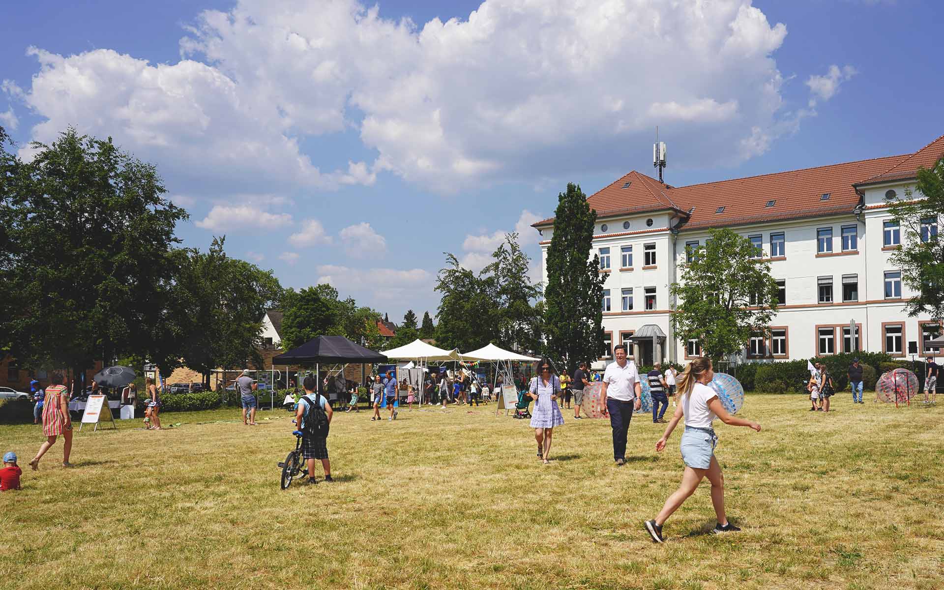 Campuswiese mit laufenden und spielenden Kindern und Erwachsenen und Ständen und Gebäude im Hintergrund