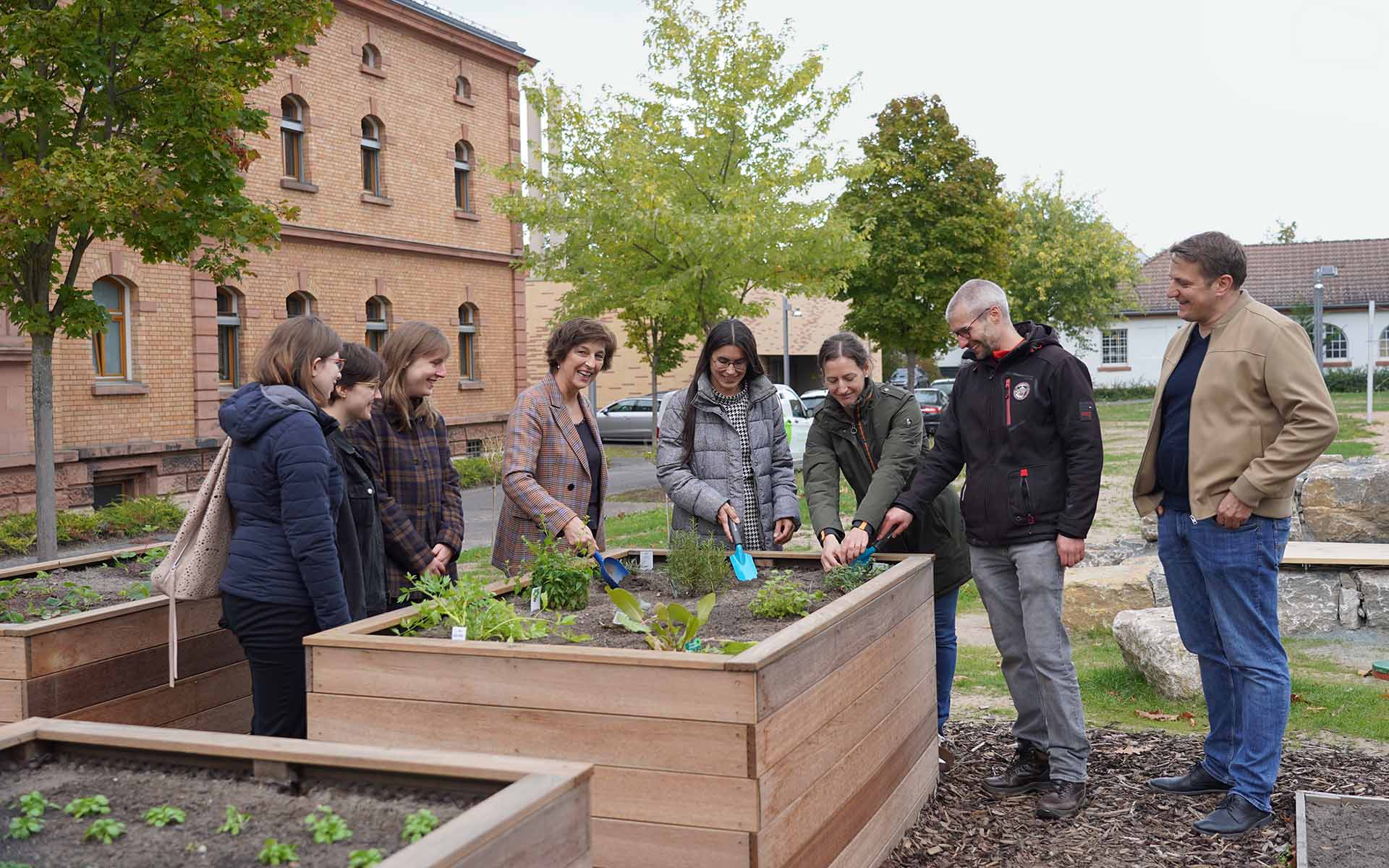 Eine Gruppe von Personen, in der Mitte die Präsidentin und die Kanzlerin stehen um ein Hochbeet mit Kräutern. Die Kanzlerin und die Präsidentin sowie die Nachhaltigkeitsbeauftragte pflanzen mit kleinen Schaufeln Kräuter ein.