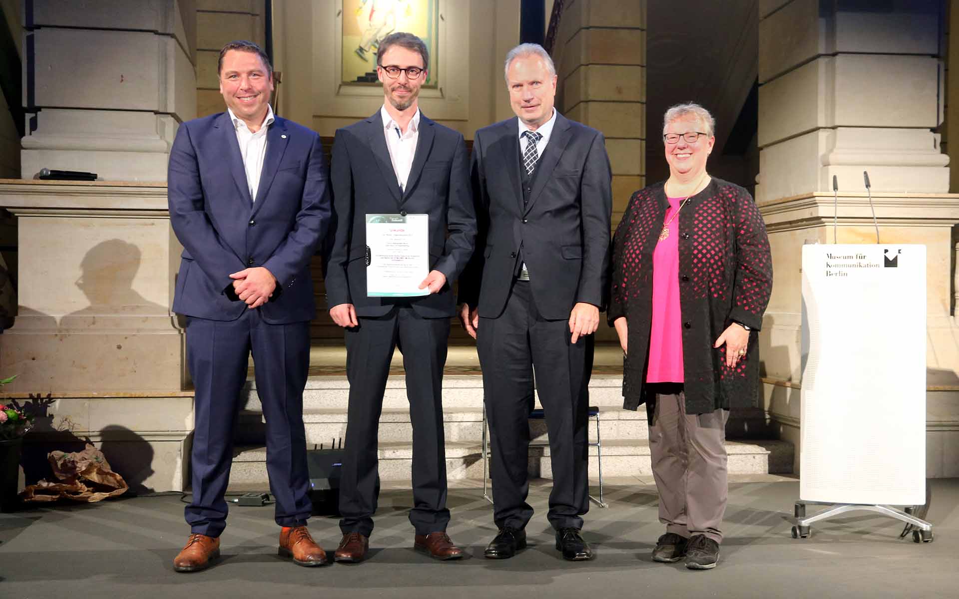 Der Preisträger hält lächelnd seine Urkunde in den Händen. Neben im in einer Reihe stehen Professor Mann, der IfKOM-Bundesvorsitzende Heinz Leymann und Cornelia Böhmer