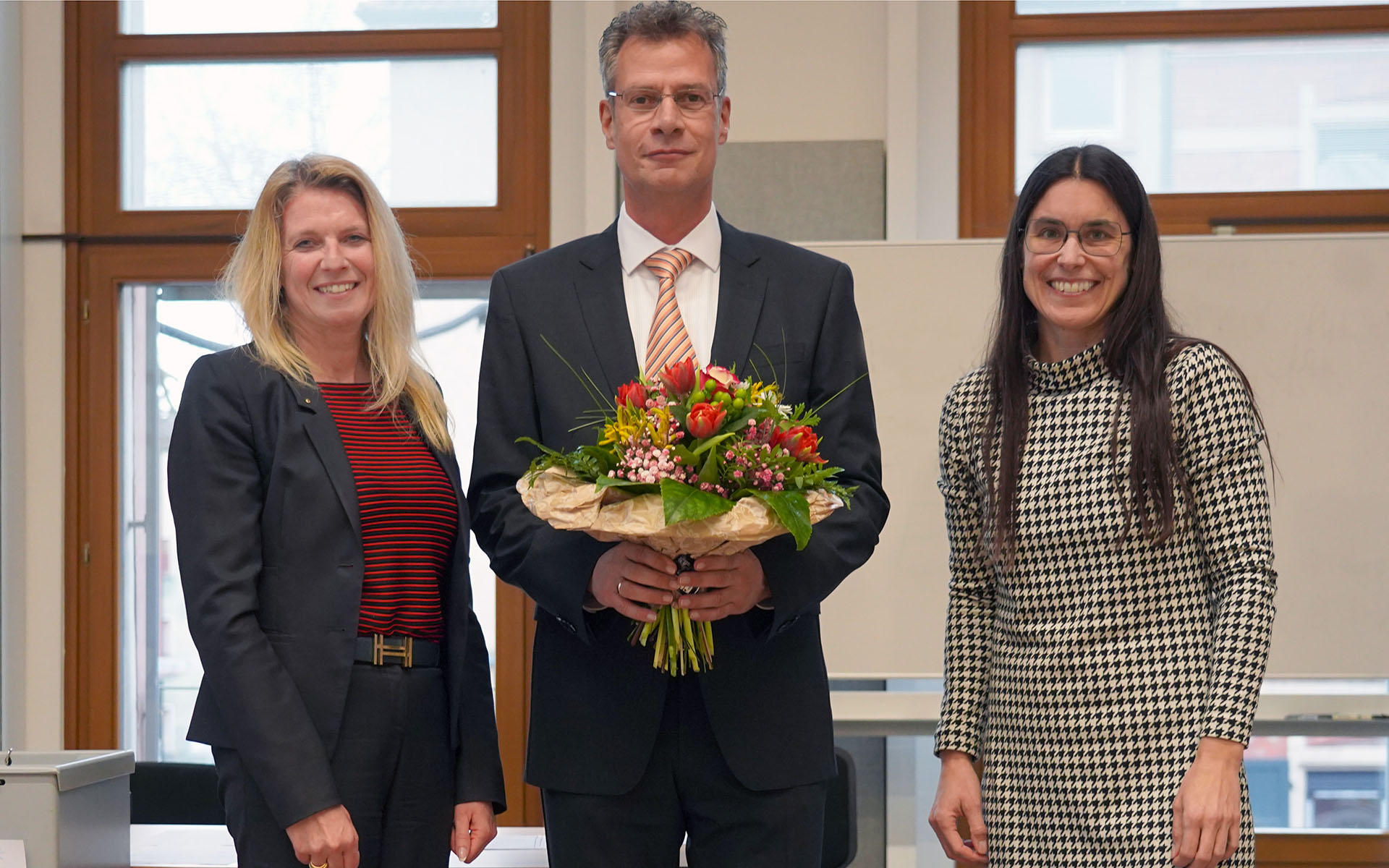 Der neue Vizepräsident steht in der Mitte auf der Bühne der Aula mit einem Blumenstrauß in der Hand, rechts neben ihm die Kanzlerin, links neben ihm die Hochschulratsvorsitzende