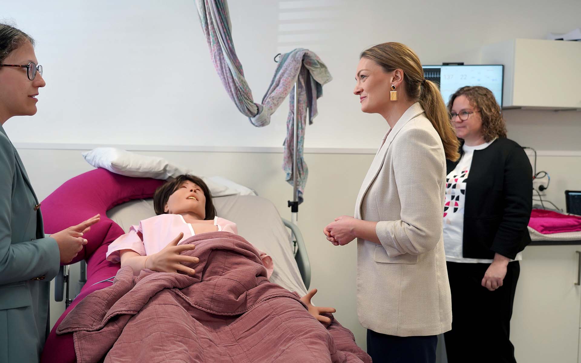 Gesundheitsministerin Judith Gerlach steht im Skills Lab vor einem Bett mit einer lebensgroßen Puppe, die eine schwangere Frau verkörpert, und spricht mit Professorin Hemma Pfeifenberger 