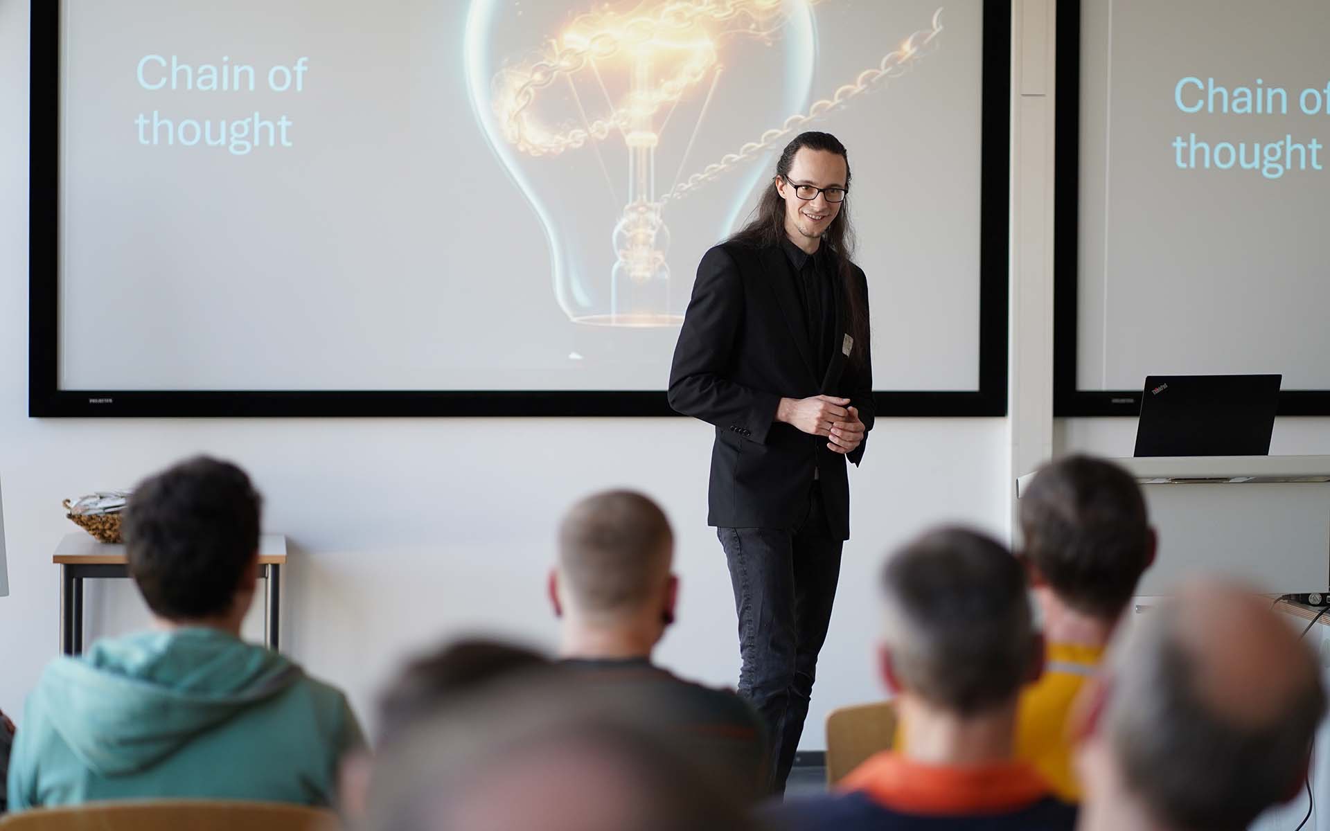 Ben Lenk-Ostendorf bei seinem Impulsvortrag mit Blick zum Publikum, im Hintergrund eine Glühbirne auf der Leinwand mit Überschrift Chain of thought