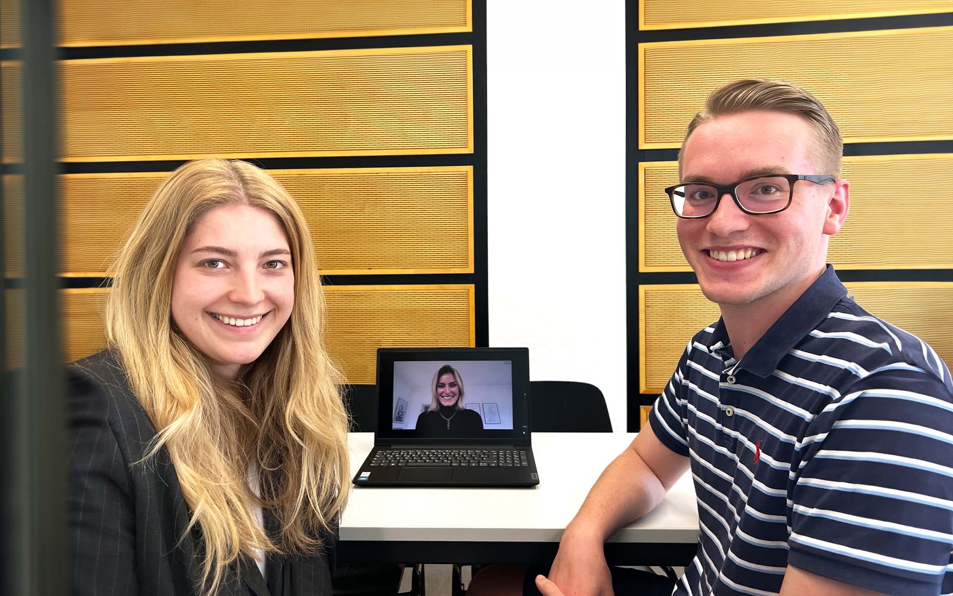 Eine Studentin und ein Student sitzen an einem Tisch. In der Mitte steht ein Laptop, auf dem gerade ein Meeting stattfindet.