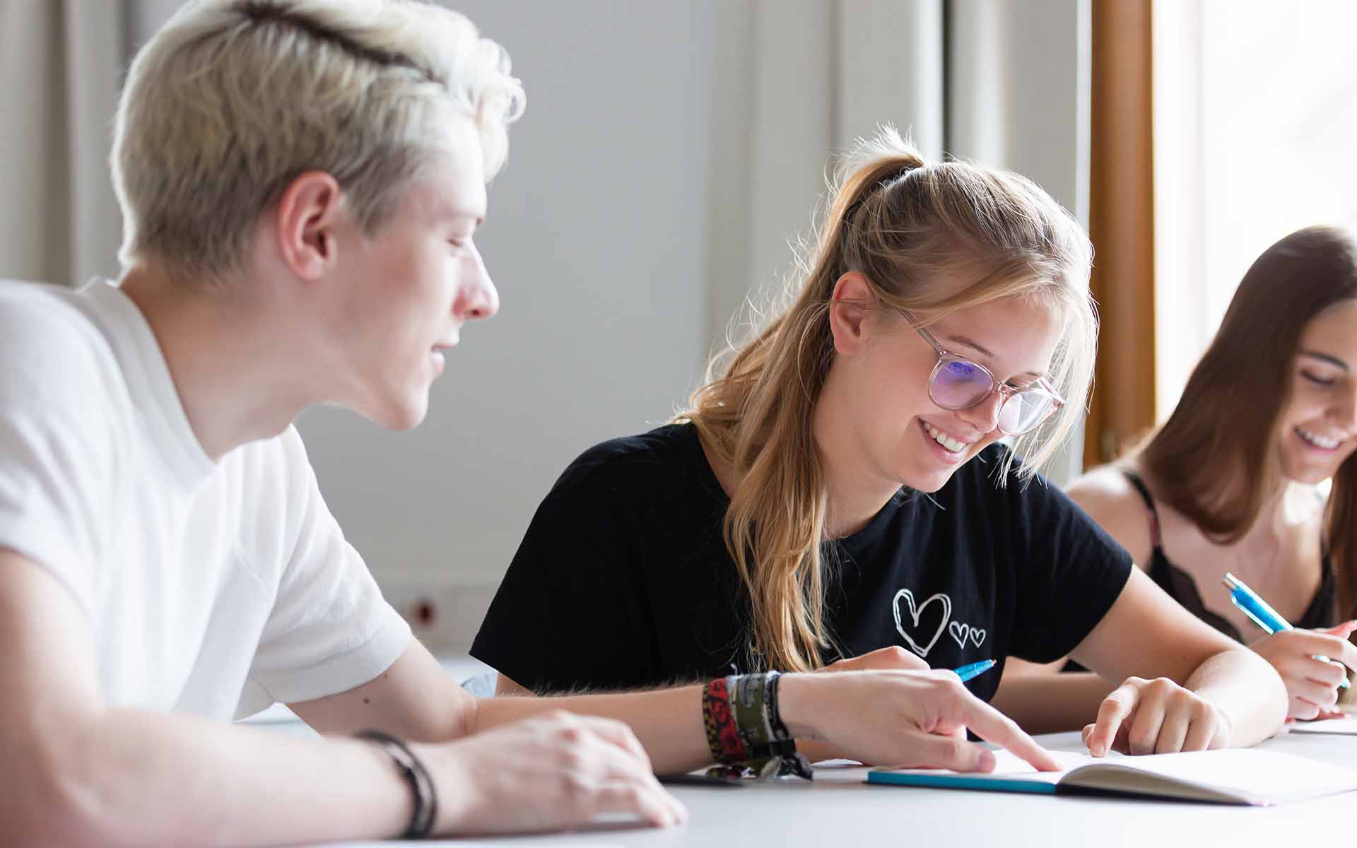 Ein Student und eine Studentin sitzen nebeneinander an einem Tisch und schauen gemeinsam in ein Buch.