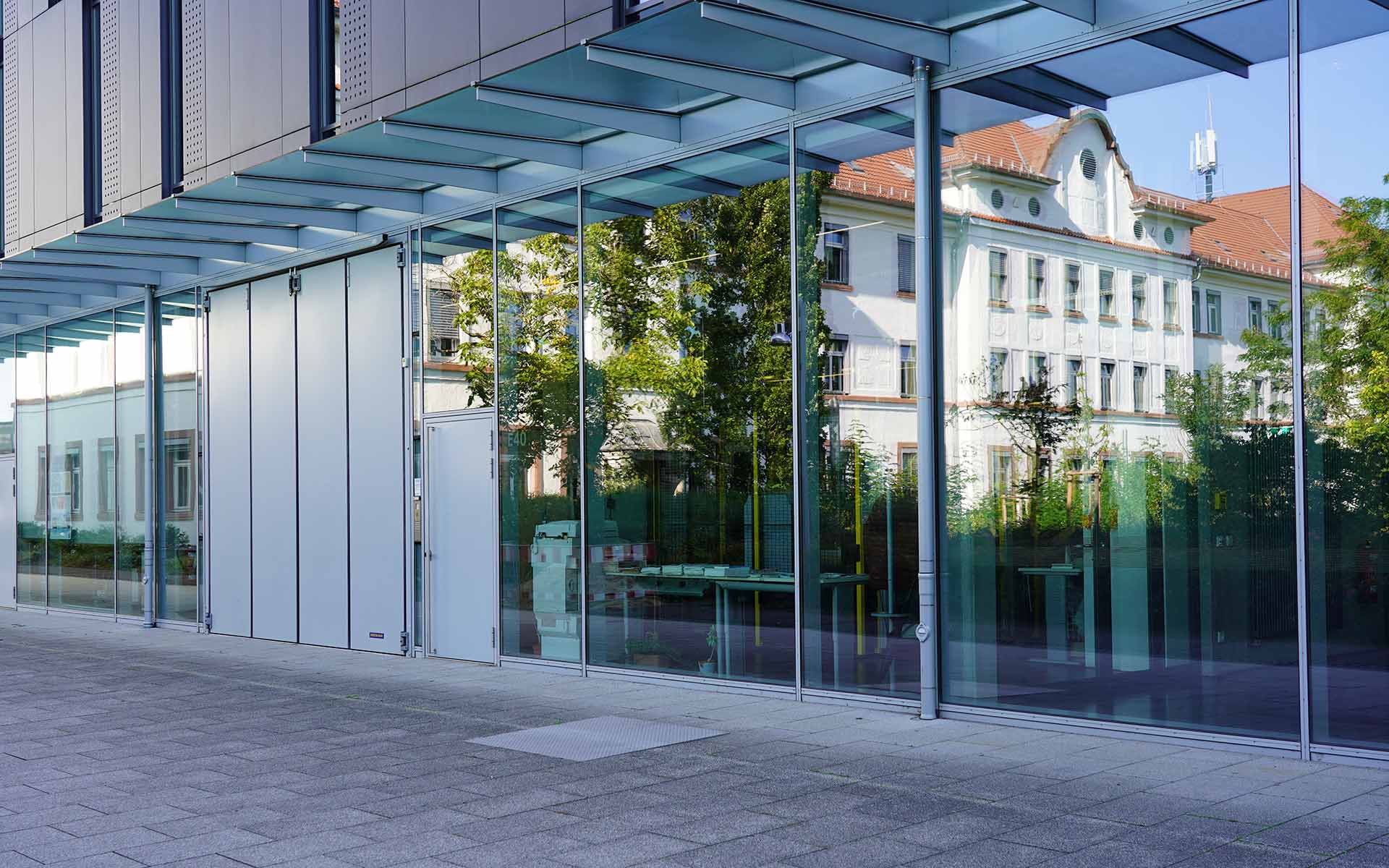 Weißes Gebäude der Hochschule mit rotem Ziegeldach spiegelt sich in der verglasten Fassade des modernen grauen Hochschulgebäudes