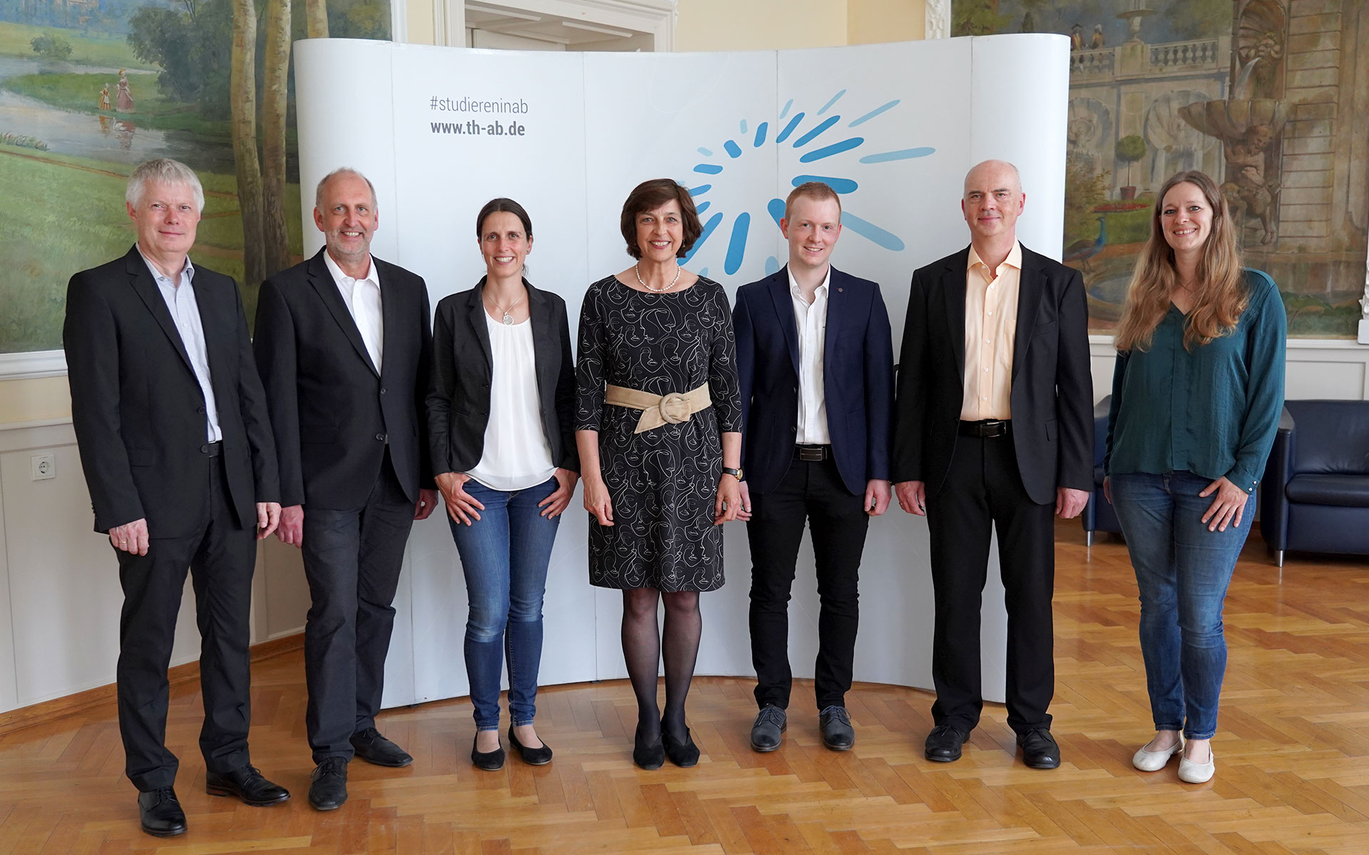 Sieben Personen stehen im Hocksaal vor einer Messewand mit Hochschullogo