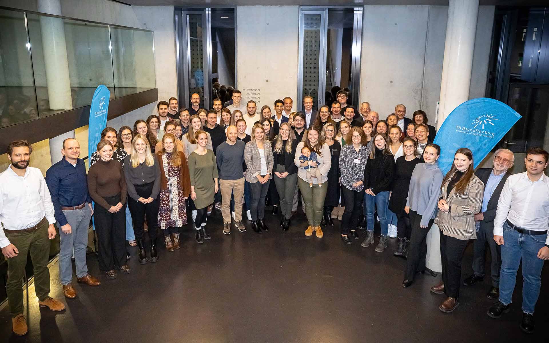 Eine Gruppe von 59 Personen steht im Flur der Hochschule im Halbkreis. Die Personen sind eingerahmt von zwei blauen Hochschul-Dropflags und lächeln in die Kamera.