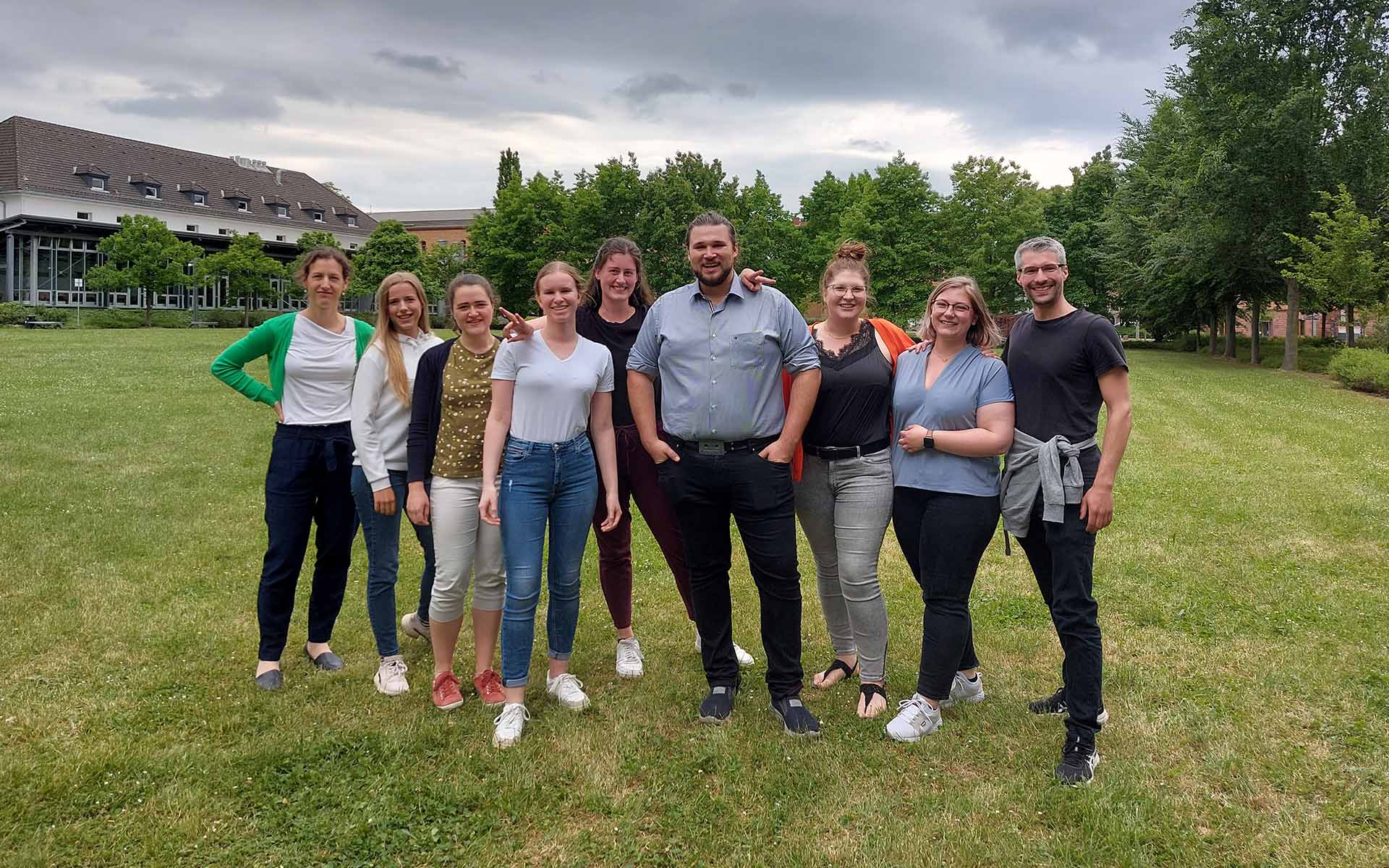 Eine Gruppe von neun Personen steht auf der grünen Campuswiese vor grünen Bäumen und dem Gebäude der Mensa bei leicht bewölktem Himmel