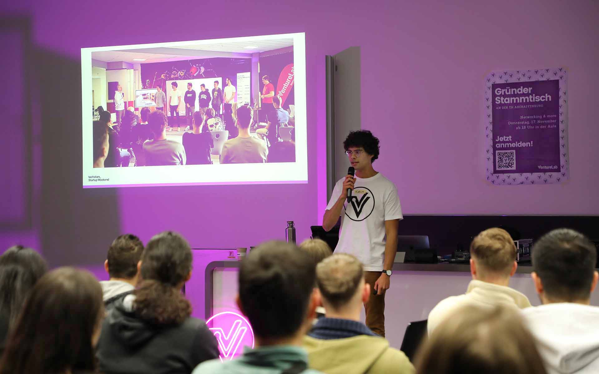 Ein junger Mann mit dunklen Locken und Brille steht vor einer pink beleuchteten Leinwand und spricht vor einer Gruppe sitzender Personen ins Mikrophon